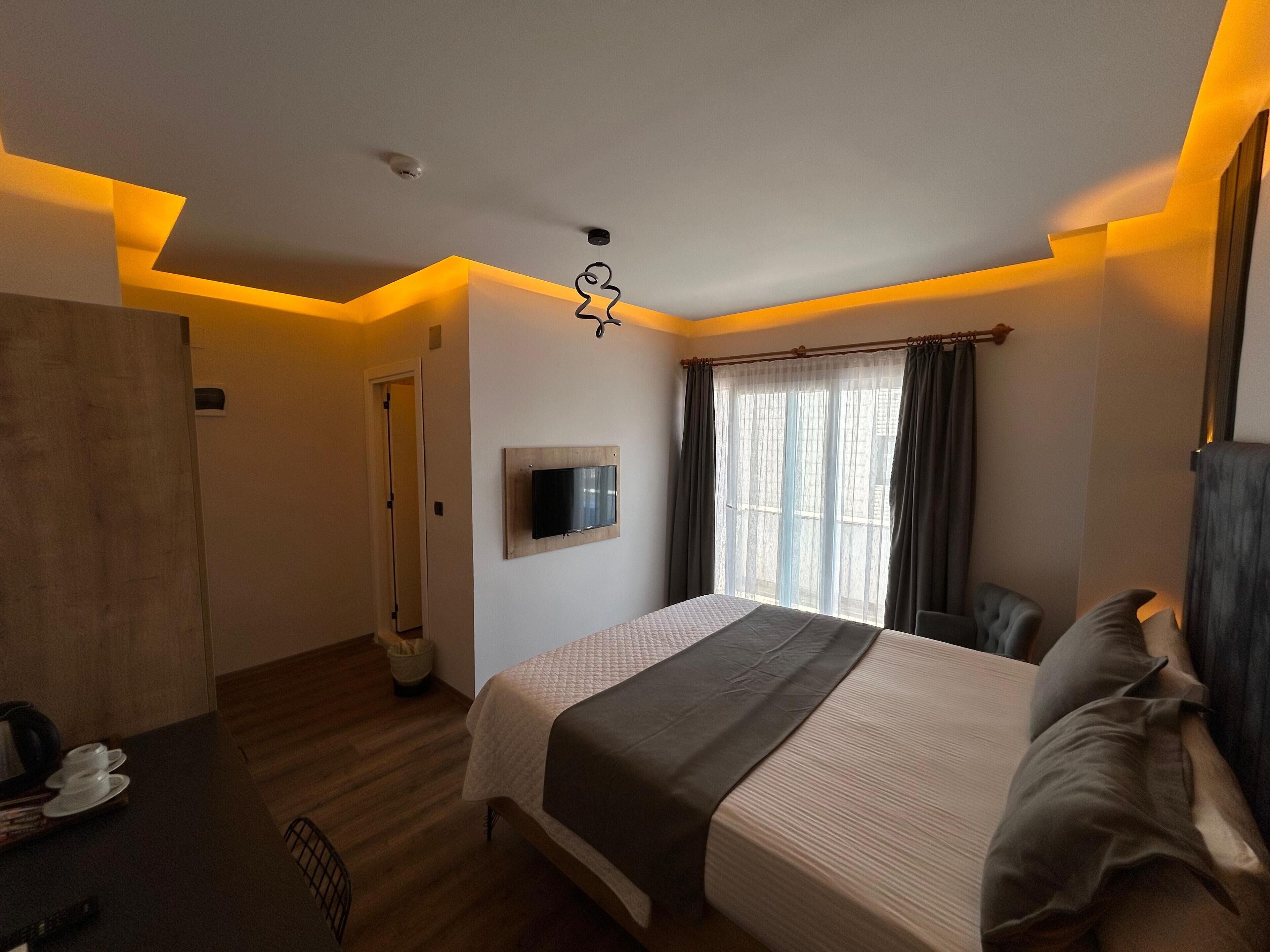 Ein Hotelzimmer mit einem Bett, einem an der Wand befestigten Fernseher, einem Fenster mit Vorhängen und einem Holzboden.