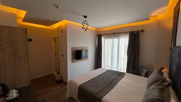Ein Hotelzimmer mit einem Bett, einem an der Wand befestigten Fernseher, einem Fenster mit Vorhängen und einem Holzboden.