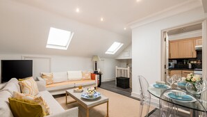 A modern living room with a sofa, coffee table, and dining area with clear glass chairs and a glass table. There is a flat-screen TV mounted on the wall, a ceiling fan, and skylights. - Luxury Notting Hill 3 bedroom, 2 Bathroom Duplex Apartment (London)