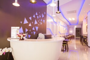 Una moderna zona de recepción con mostrador blanco, pared decorativa con diseños geométricos y un área de estar con mesas y sillas.