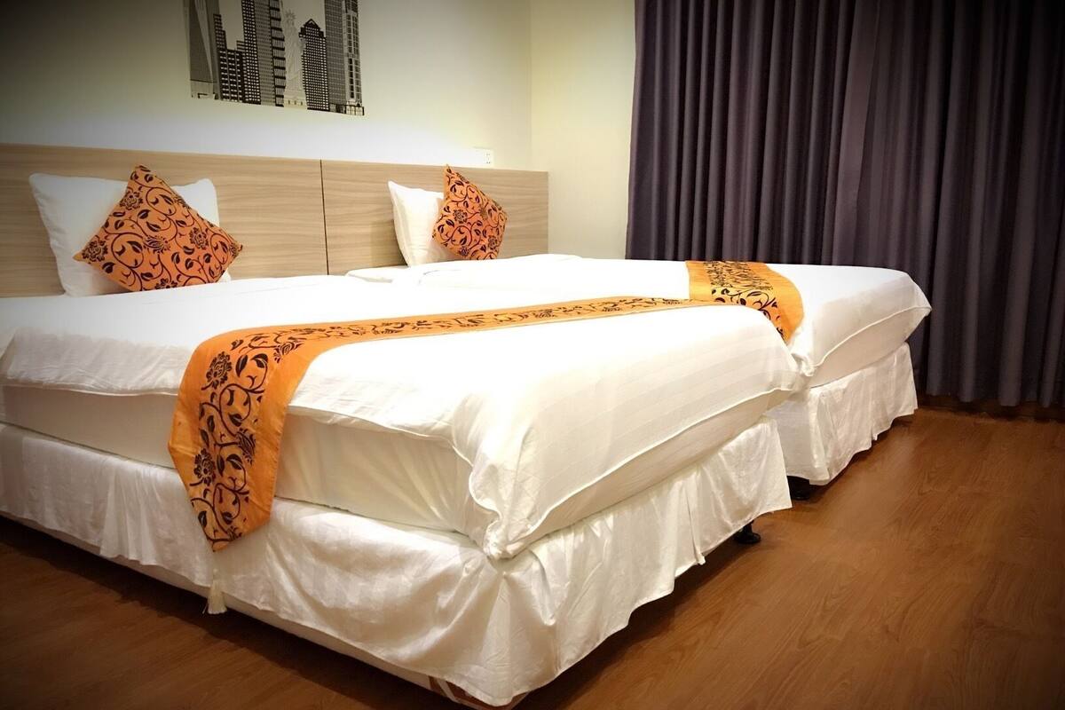 A double bed with white bedding and orange decorative pillows, a wooden headboard, and a dark curtain.