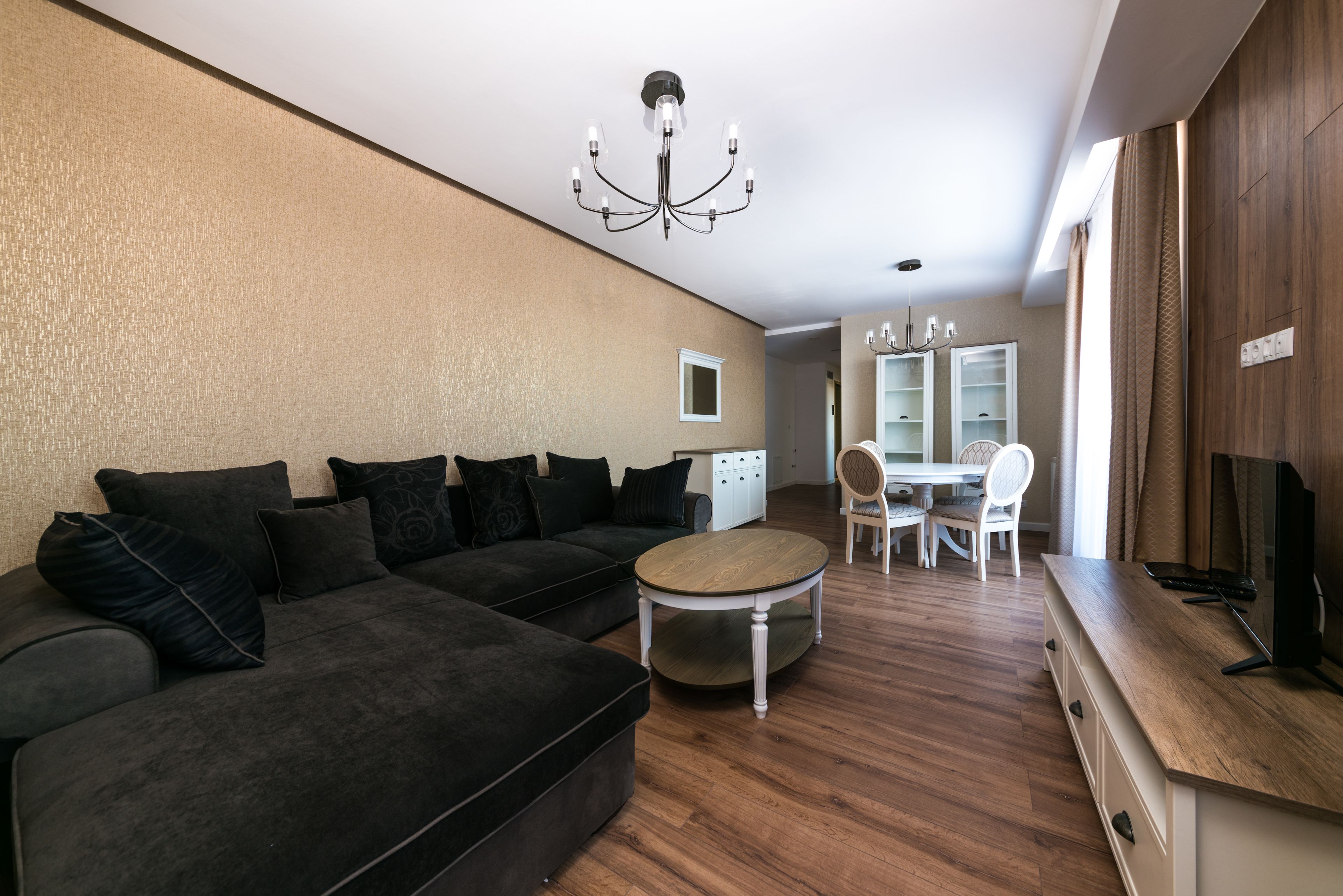 A modern living room with a sectional sofa, a round coffee table, a dining area with a white table and chairs, and a TV on a wooden cabinet.