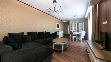 A modern living room with a sectional sofa, a round coffee table, a dining area with a white table and chairs, and a TV on a wooden cabinet.