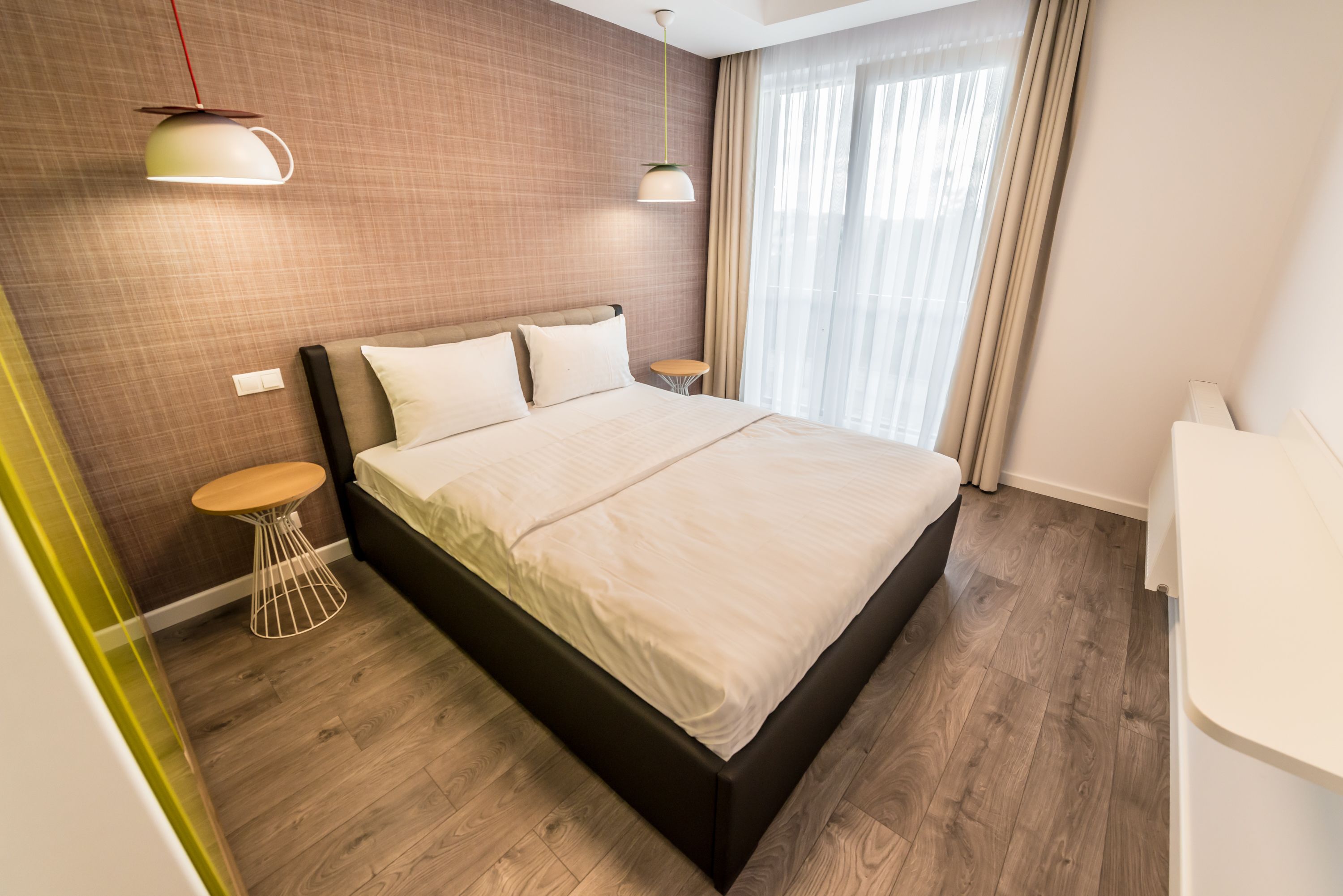 Ein modernes Schlafzimmer mit einem großen Bett, zwei Nachttischen, einer Pendelleuchte und Blick ins Freie durch transparente Vorhänge.
