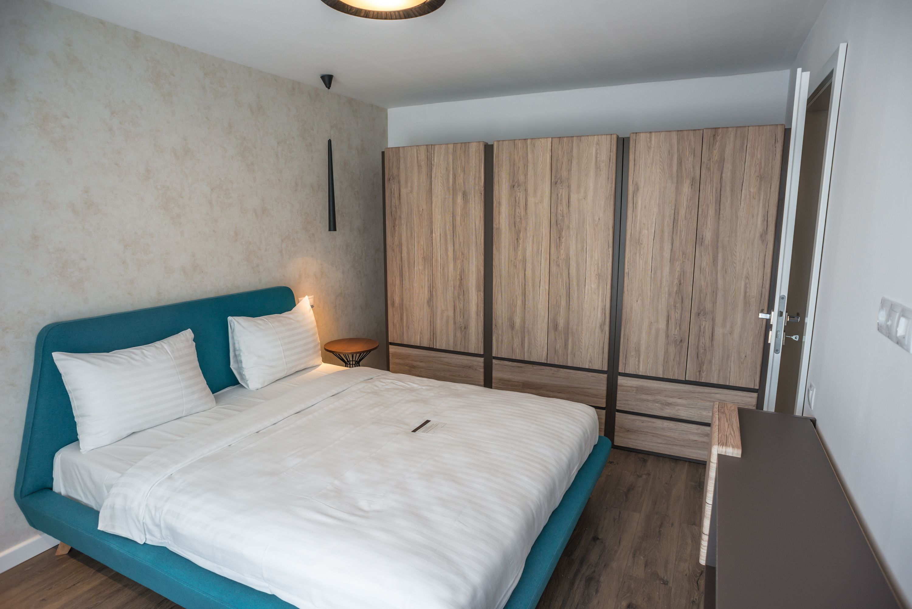 A bedroom with a teal bed, white bedding, a wooden wardrobe, and a small bedside table with a lamp.