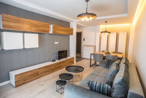 A modern living room with a grey sofa, a wooden console table, and a TV. There is a dining area with a table and chairs.