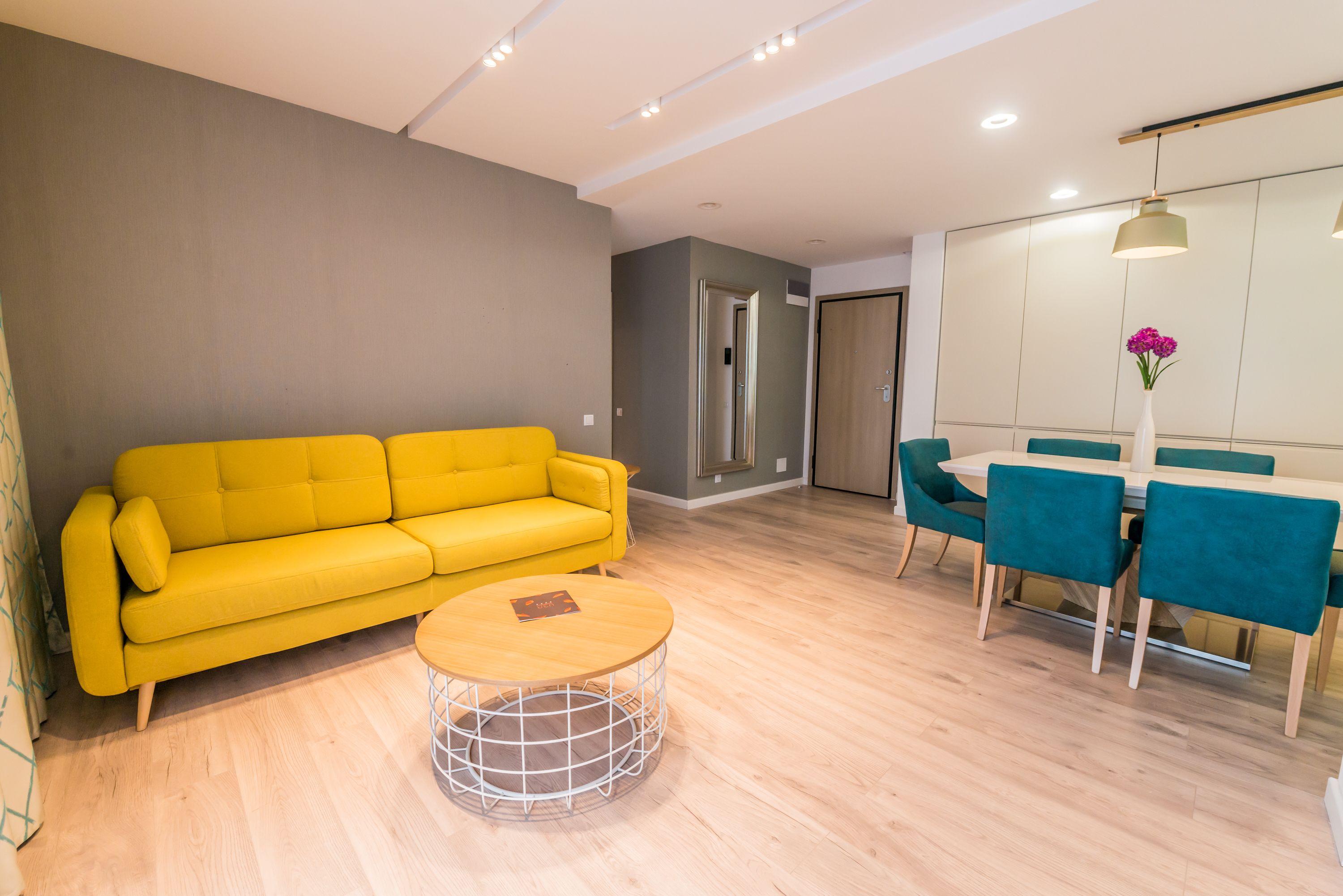 A modern living room with a yellow sofa, a round coffee table, teal chairs, and a dining table.
