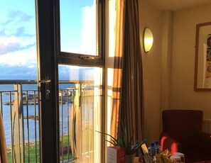 A room with a large window overlooking a body of water, a red chair, a potted plant, and a wall-mounted light fixture.