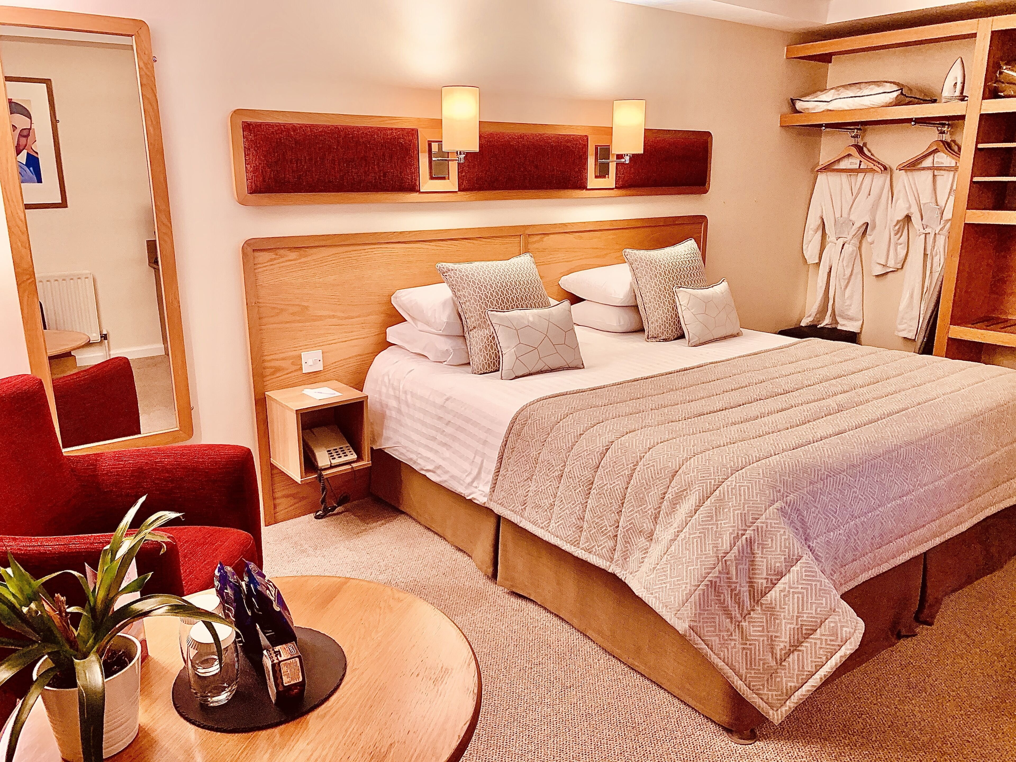 Una habitación de hotel con una cama grande, una mesita de noche con lámpara, un sillón rojo, una pequeña mesa con una planta y una botella, y un armario con ropa.