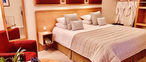 Una habitación de hotel con una cama grande, una mesita de noche con lámpara, un sillón rojo, una pequeña mesa con una planta y una botella, y un armario con ropa.