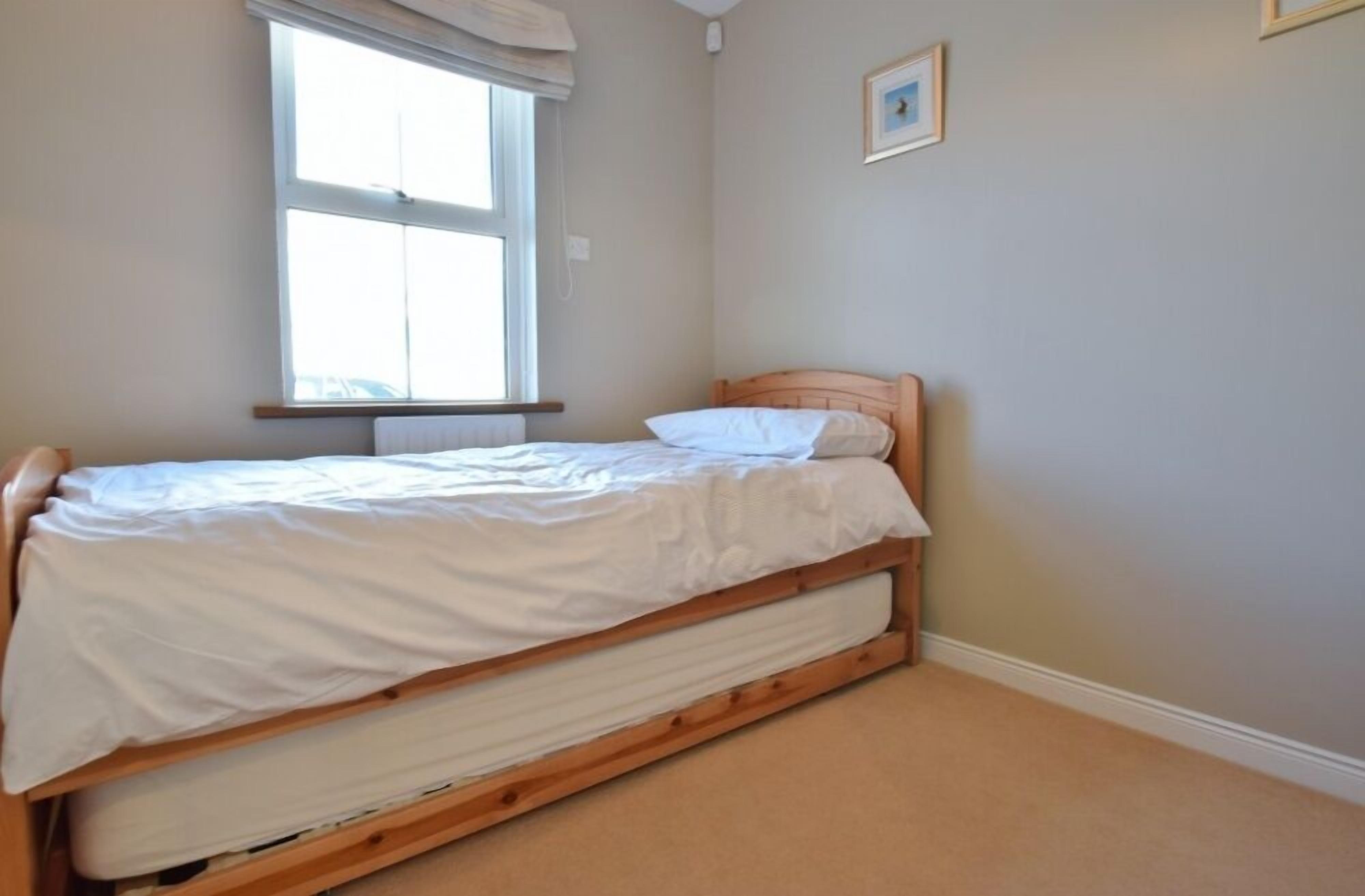 A single bed with a wooden frame and white bedding in a room with beige walls and a window with white curtains.