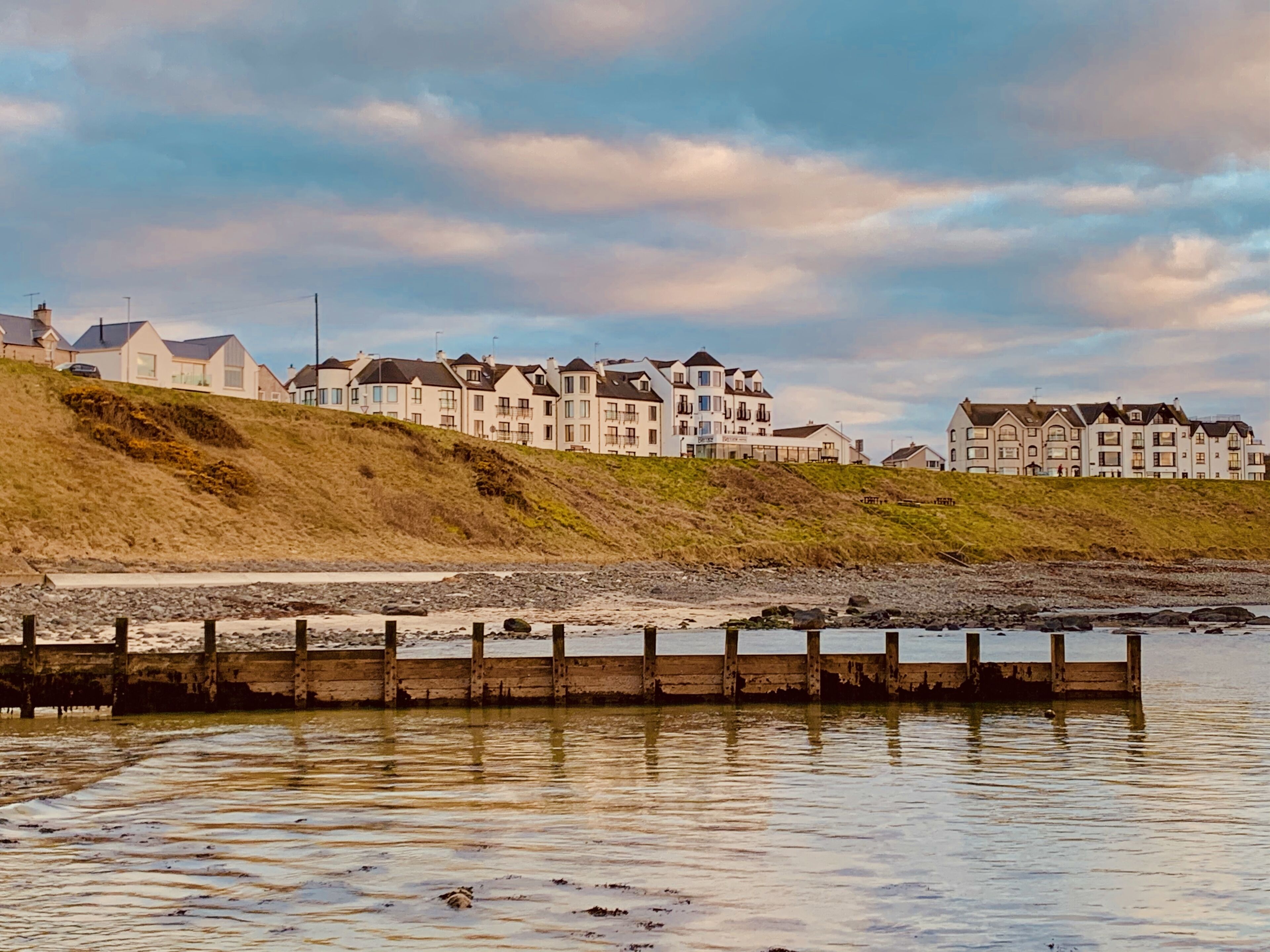 A coastal residential area with multi-story houses, a wooden jetty, and a grassy hillside.