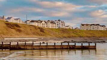 A coastal residential area with multi-story houses, a wooden jetty, and a grassy hillside.