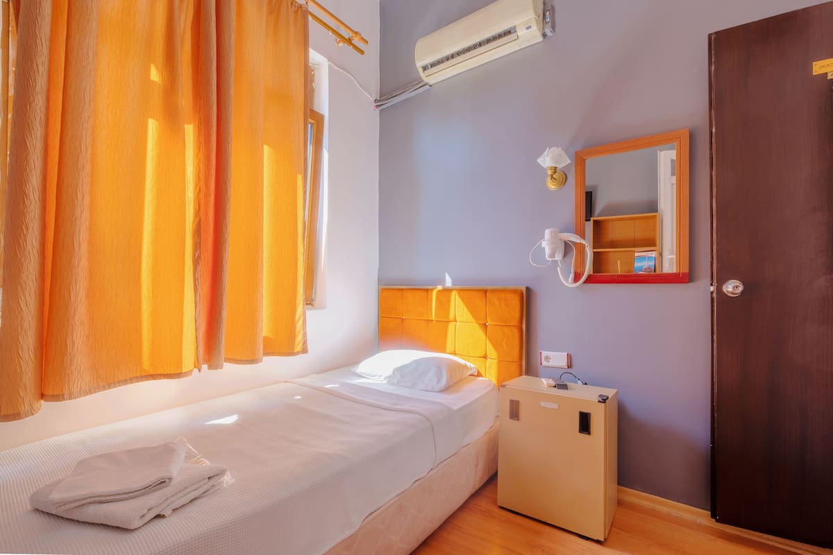 Un lit simple avec un matelas et des oreillers blancs, un appareil de climatisation au-dessus, une petite table de chevet avec une lampe et une armoire en bois.