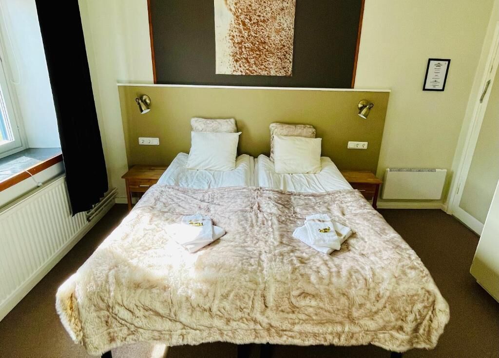 Uma cama de casal com um cobertor bege fofinho, duas mesas de cabeceira, uma janela com cortinas e uma obra de arte pendurada na parede.