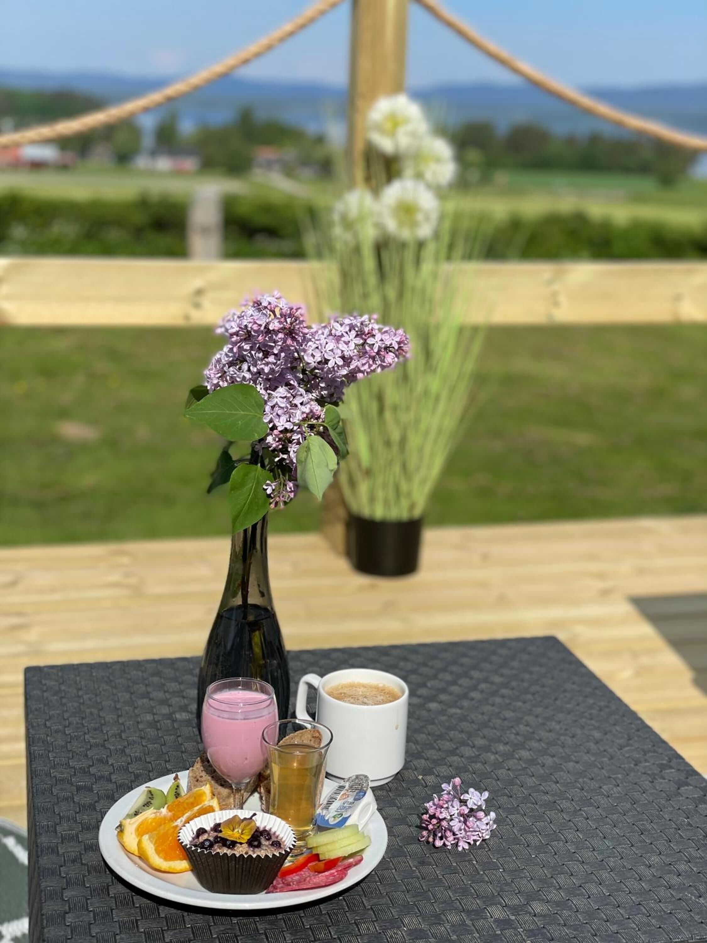 A table with a meal, a drink, and a flower arrangement on a deck with a view of a field and mountains.