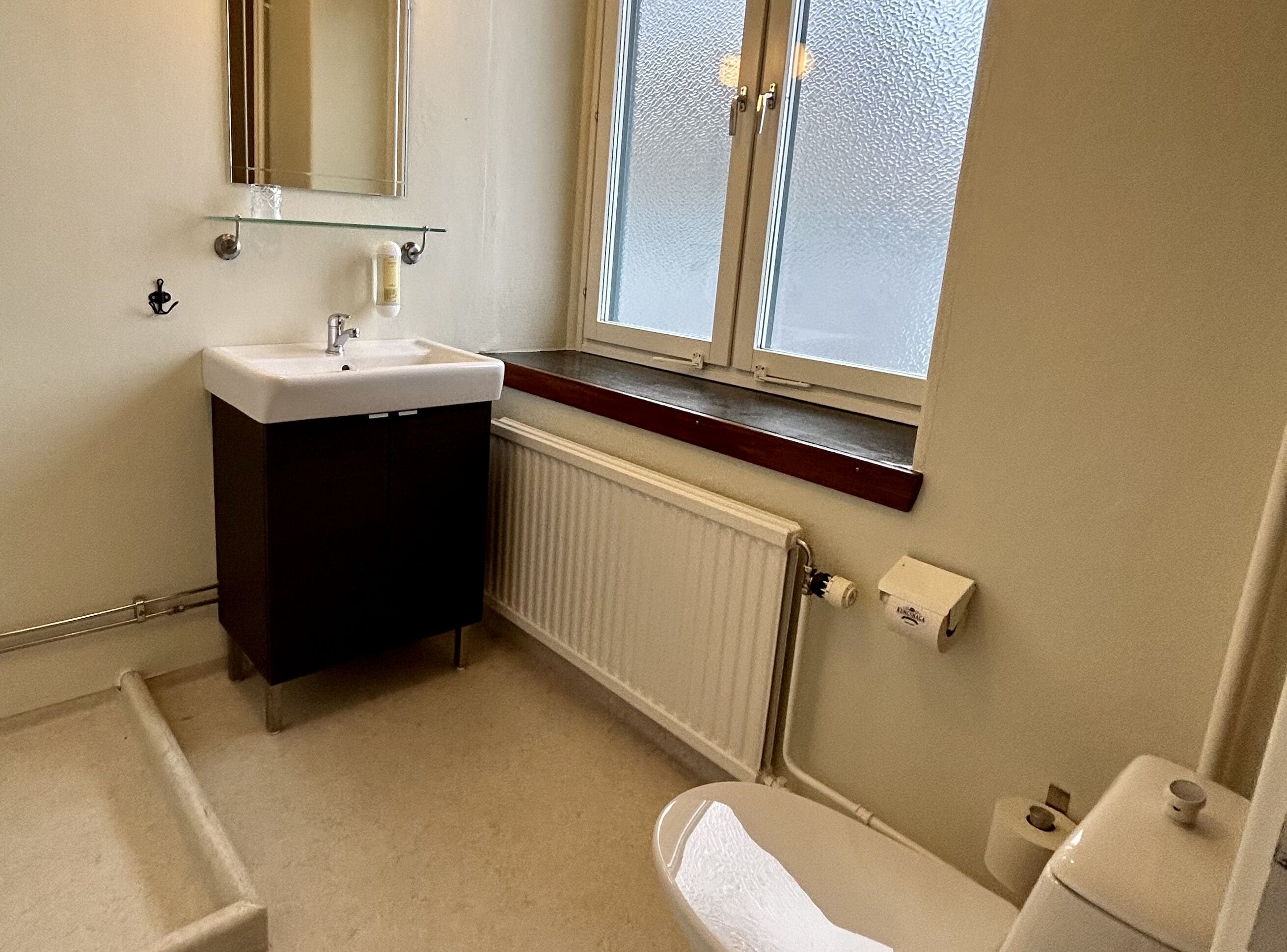 A small bathroom with a white sink, dark cabinet, window, radiator, and toilet.