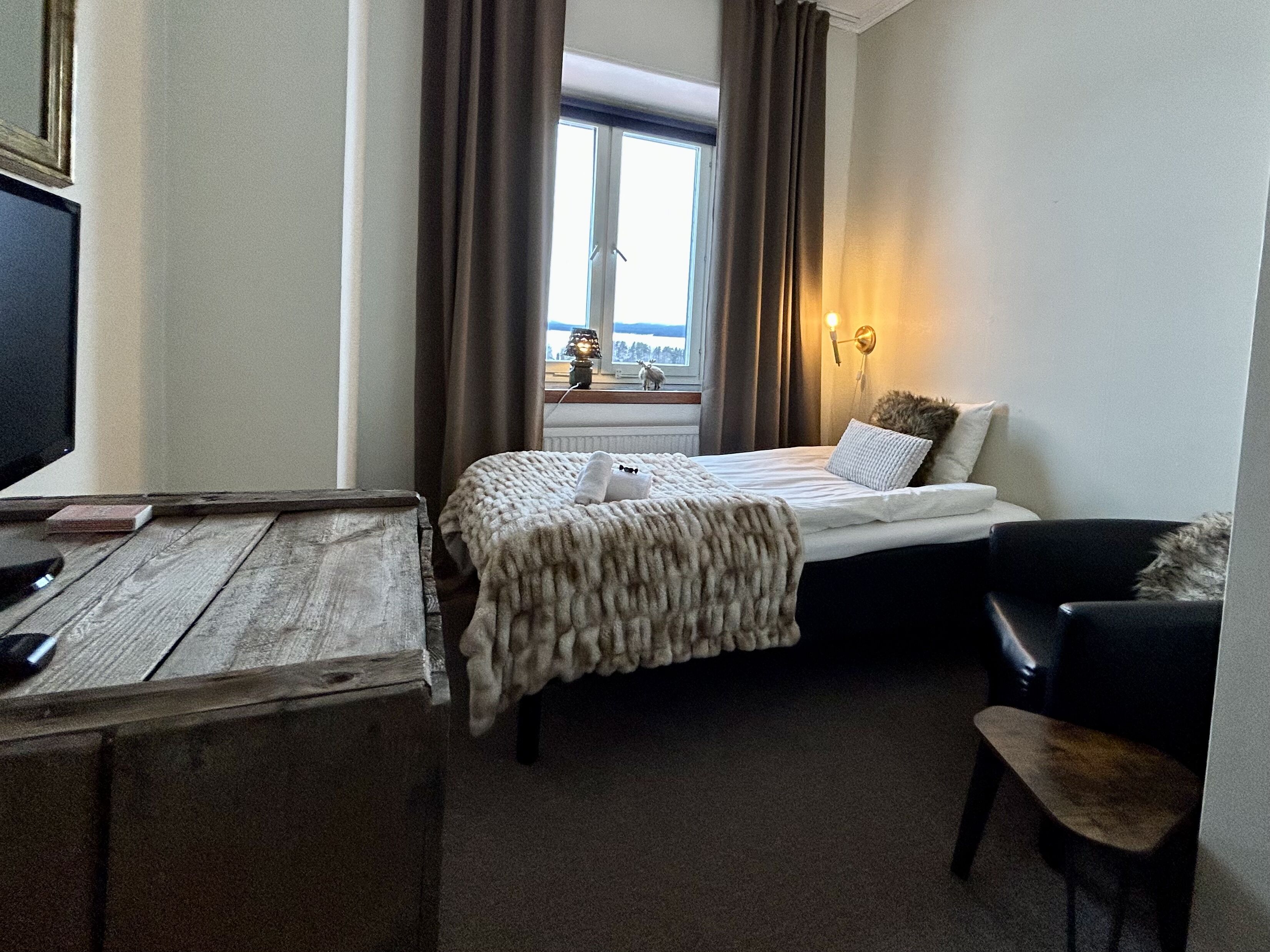 Quarto de hotel com cama, escrivaninha, cadeira e janela com cortinas.