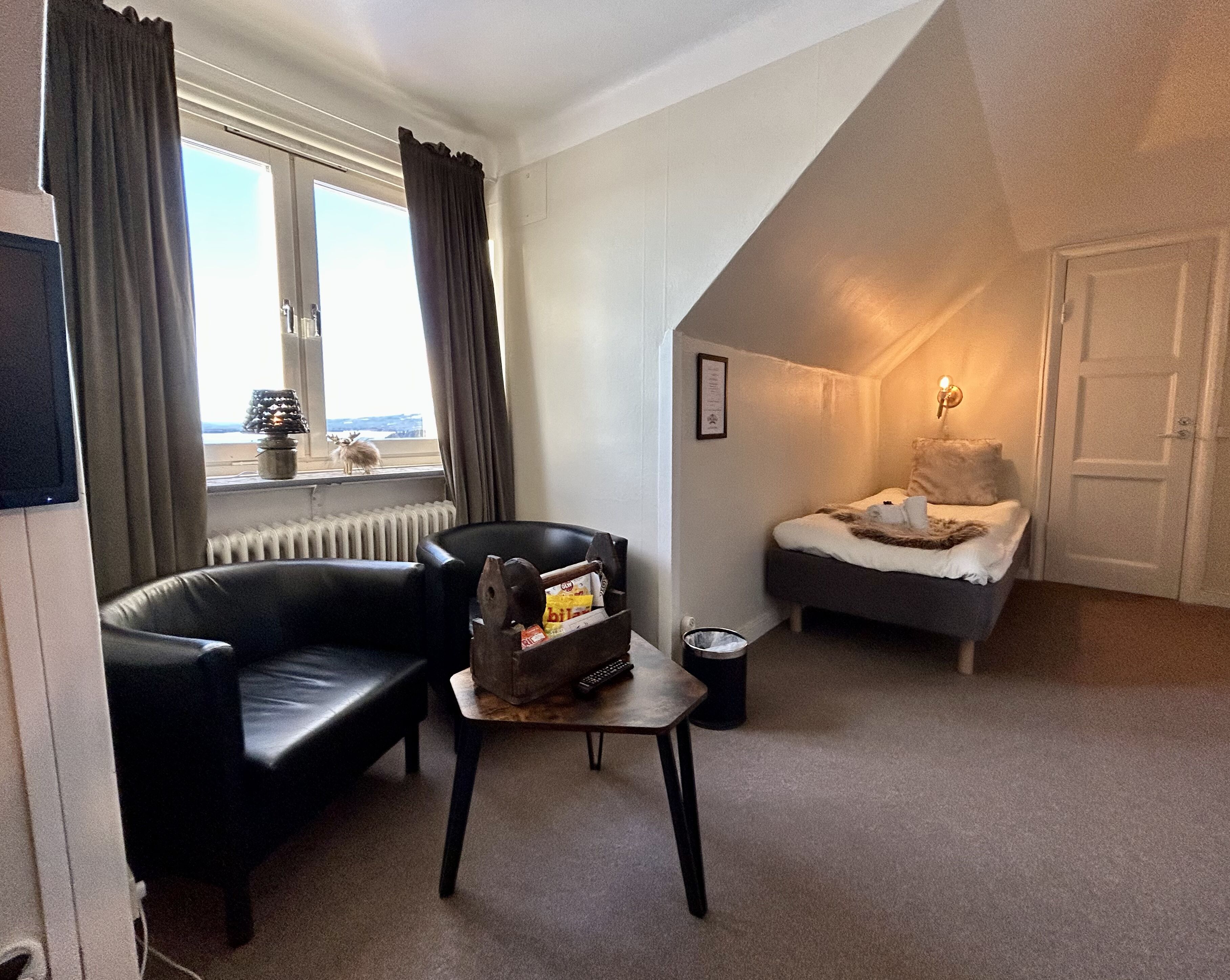 A compact room with a black leather sofa, a small wooden table, a bed with a white blanket, a radiator, and a window with curtains.
