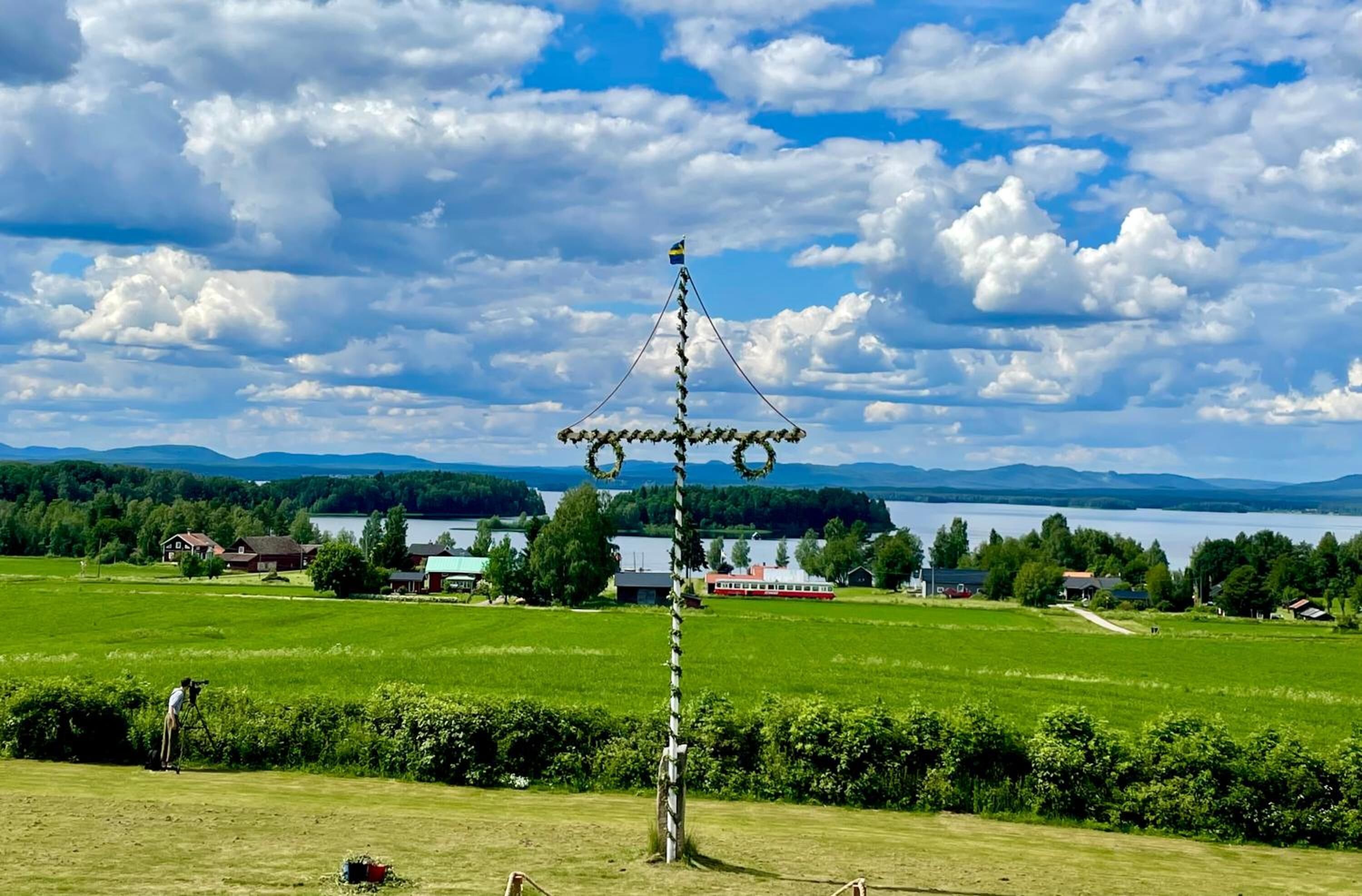 A rural landscape with a traditional wooden structure in the foreground, surrounded by green fields and houses in the background.