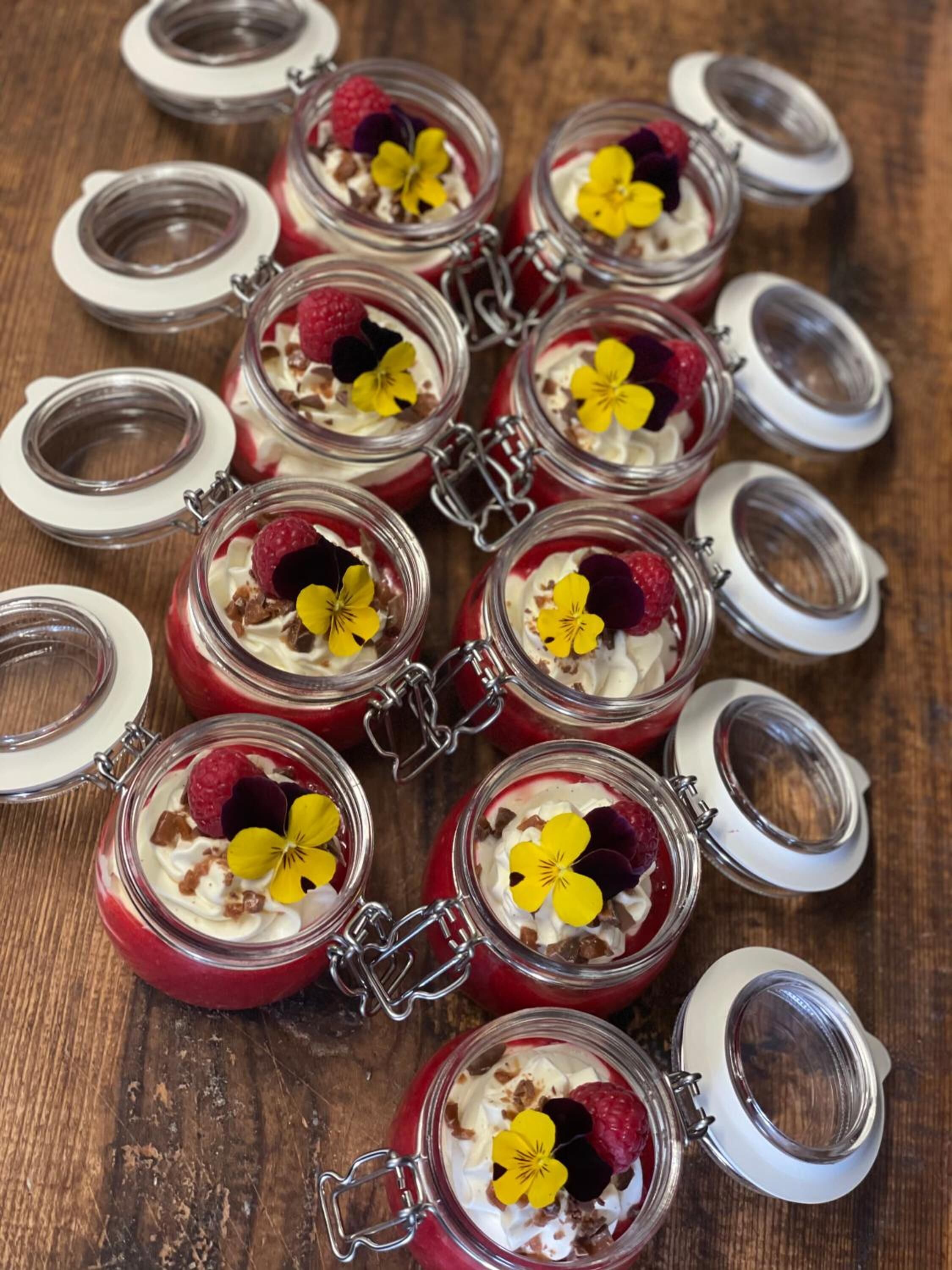 Multiple glass jars with metal clasps, each containing a dessert with whipped cream and berries, and a yellow flower on top.