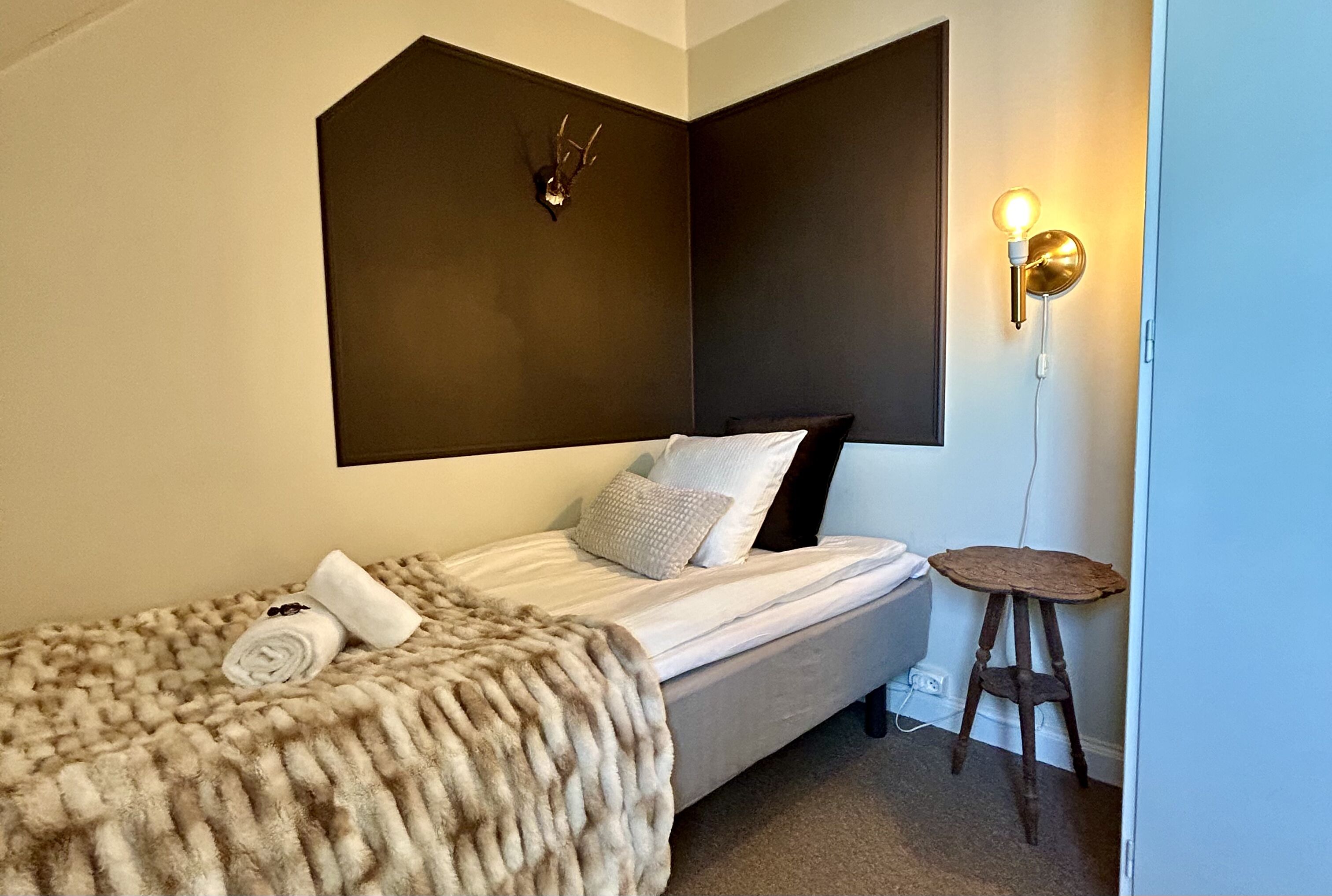 Een slaapkamer met een bed, een bontdeken, een aan de muur bevestigd hertenhoofd, een wandlamp en een klein houten krukje.