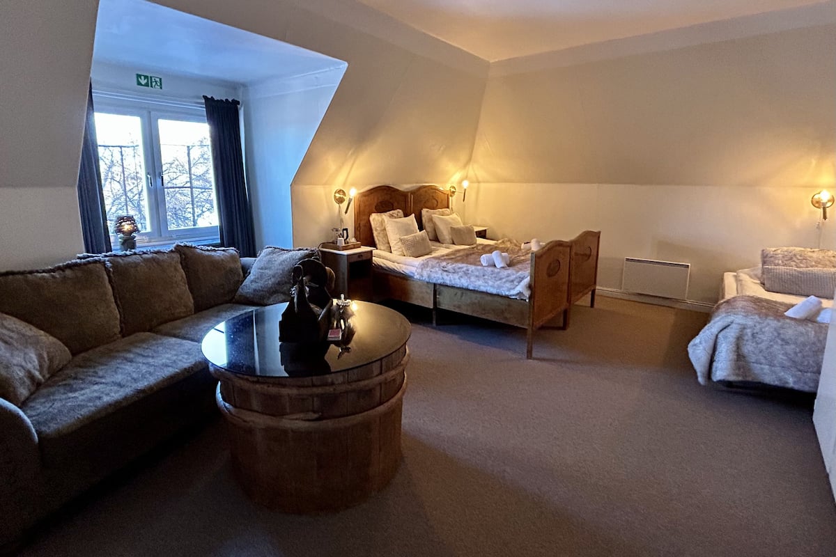 Ett hotellrum med en soffa, ett runt soffbord i trä, en säng med en sänggavel och ett litet nattduksbord.