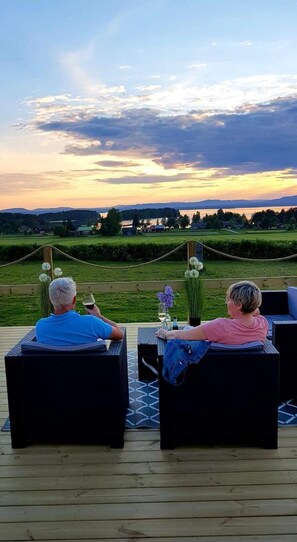 Duas pessoas sentadas em cadeiras de um pátio ao ar livre, apreciando a vista de um lago e colinas ao longe durante o pôr do sol.