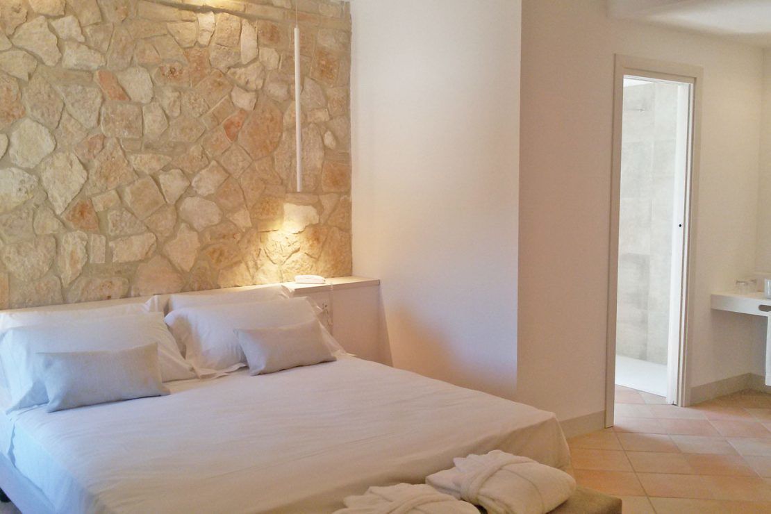 A bedroom with a stone wall, a bed with white linens, and a bathroom visible through an open door.