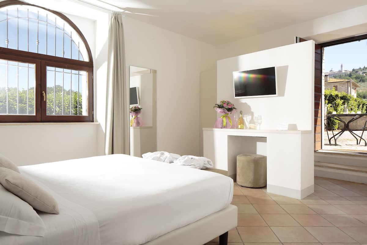 A hotel room with a bed, a TV, a desk with a mirror, and a balcony with a view of the city.