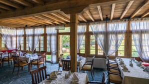 Een restaurant met houten balken en grote ramen met lichte gordijnen. Tafels zijn gedekt met witte tafelkleden en servetten.