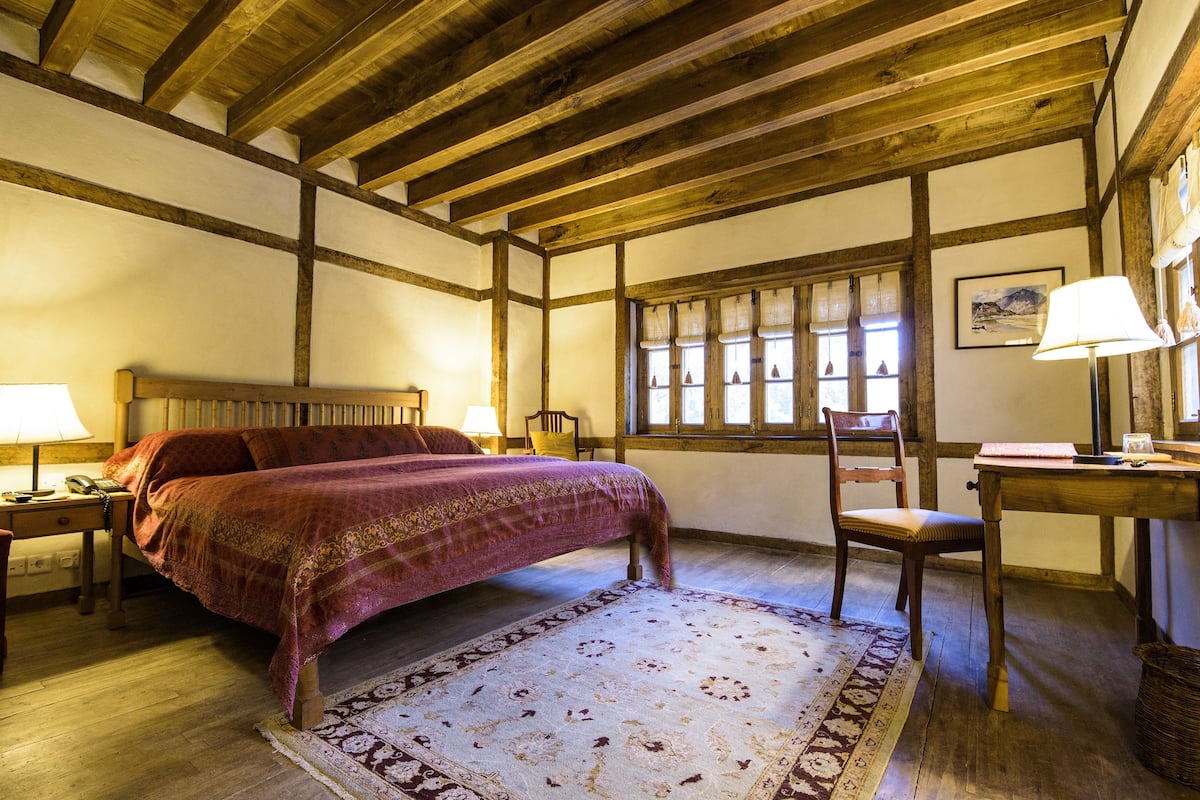 Une chambre avec un plafond en bois, un lit recouvert d’une couverture rouge, une chaise, un bureau avec une lampe et une fenêtre avec des rideaux.