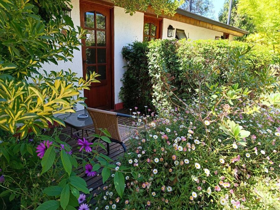 Una casa con puerta de madera, un pequeño porche con mesa y silla, y un jardín con diversas flores y plantas.
