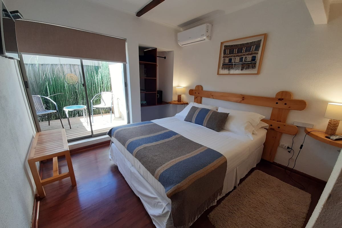 Une chambre avec un grand lit, une tête de lit en bois, une table de chevet avec une lampe et une vue sur un balcon avec des chaises.