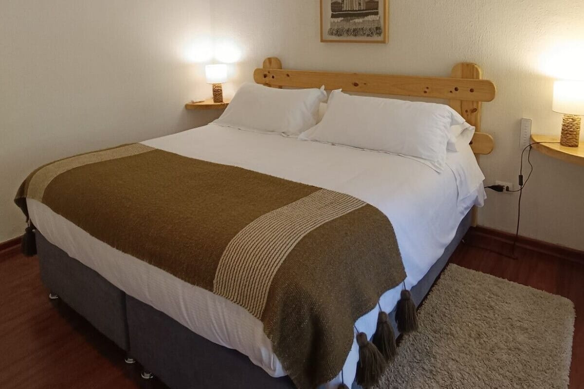 Un lit simple avec une tête de lit en bois, une literie blanche et un plaid marron. Il y a deux tables de chevet avec des lampes, un climatiseur fixé au mur et un tableau encadré au-dessus du lit.