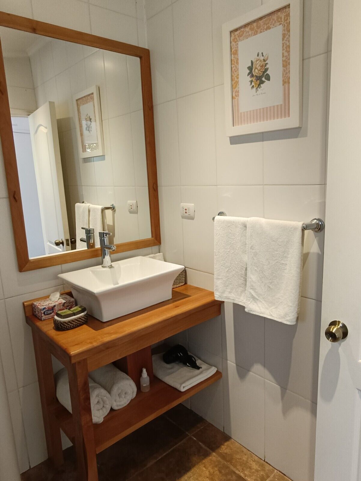 Une salle de bain avec un meuble-lavabo en bois, un lavabo blanc et un grand miroir. On y trouve des serviettes et des articles de toilette sur le meuble-lavabo, ainsi qu’un tableau encadré au mur.