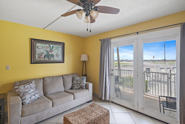 A living room with a sofa, a ceiling fan, a lamp, and a view of the outside balcony.