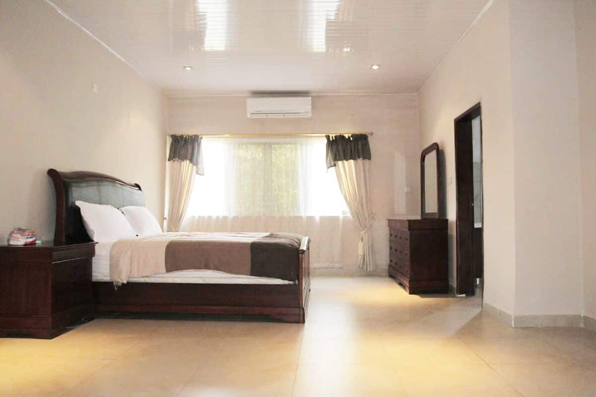 Un dormitorio con cama, mesitas de noche, cómoda, espejo y ventana con cortinas.