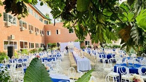 An outdoor event space with rows of tables and chairs, a brick building in the background, and large green leaves in the foreground. - Occasions (Freetown)