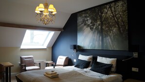Une chambre dotée d’un grand lit, d’un lustre, d’un tableau représentant des arbres, d’une lucarne et d’un coin salon avec des fauteuils.