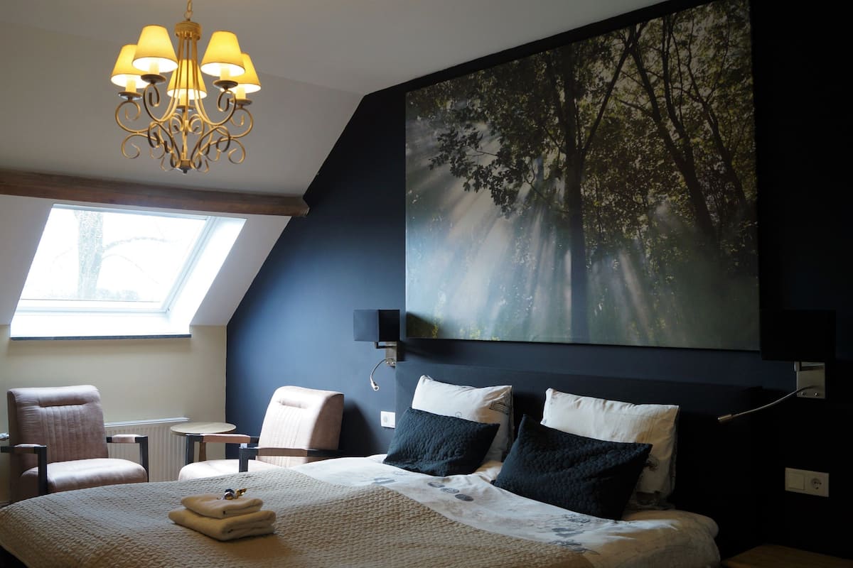 Un dormitorio con una cama grande, un candelabro, un cuadro de árboles, un tragaluz y una zona de estar con sillones.