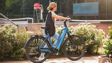 Iemand fietst op een blauwe elektrische fiets met een mandje achterop, geparkeerd nabij een hek met groen en een borduur in de achtergrond.