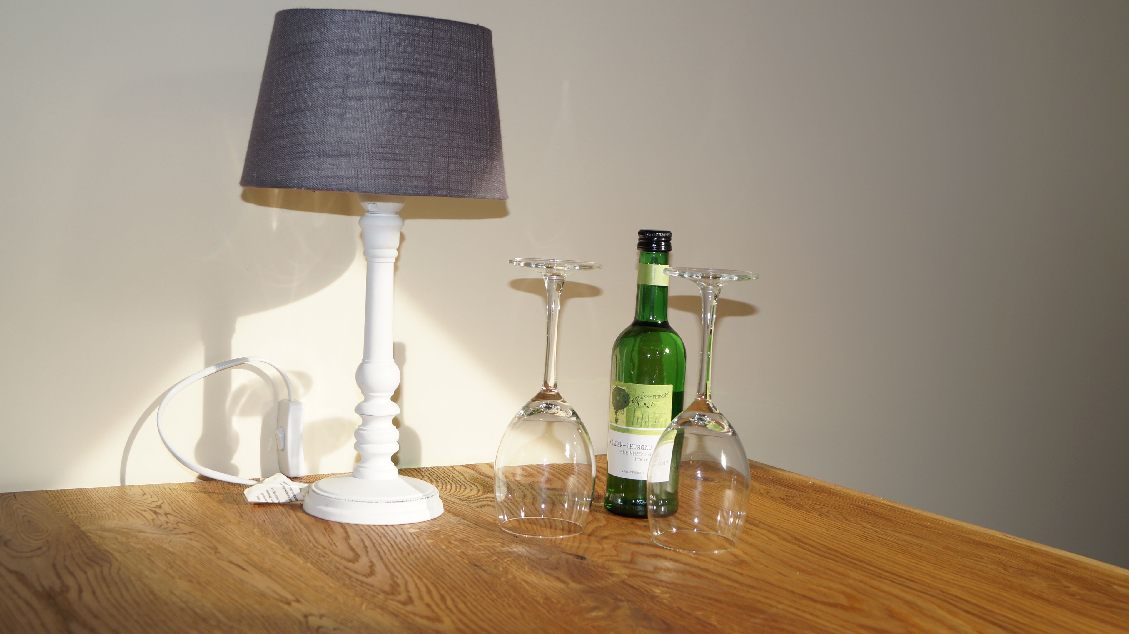 Un tavolo di legno con una lampada bianca, una bottiglia di vino verde e tre bicchieri da vino.