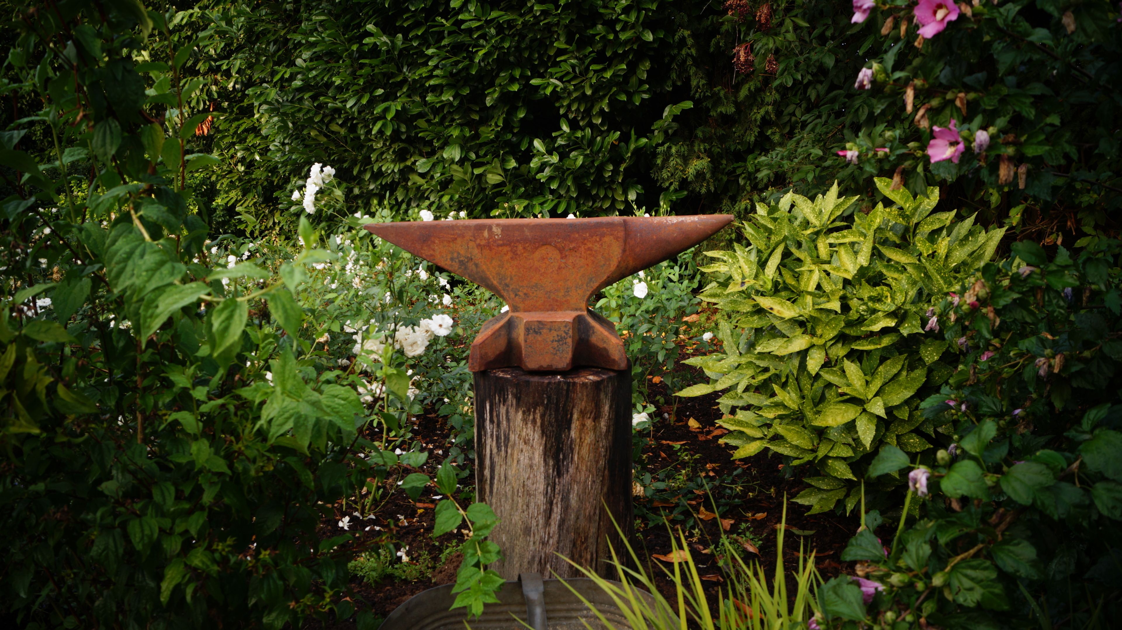 Una fontana rustica con una vasca in metallo, circondata da vegetazione lussureggiante e piante fiorite.