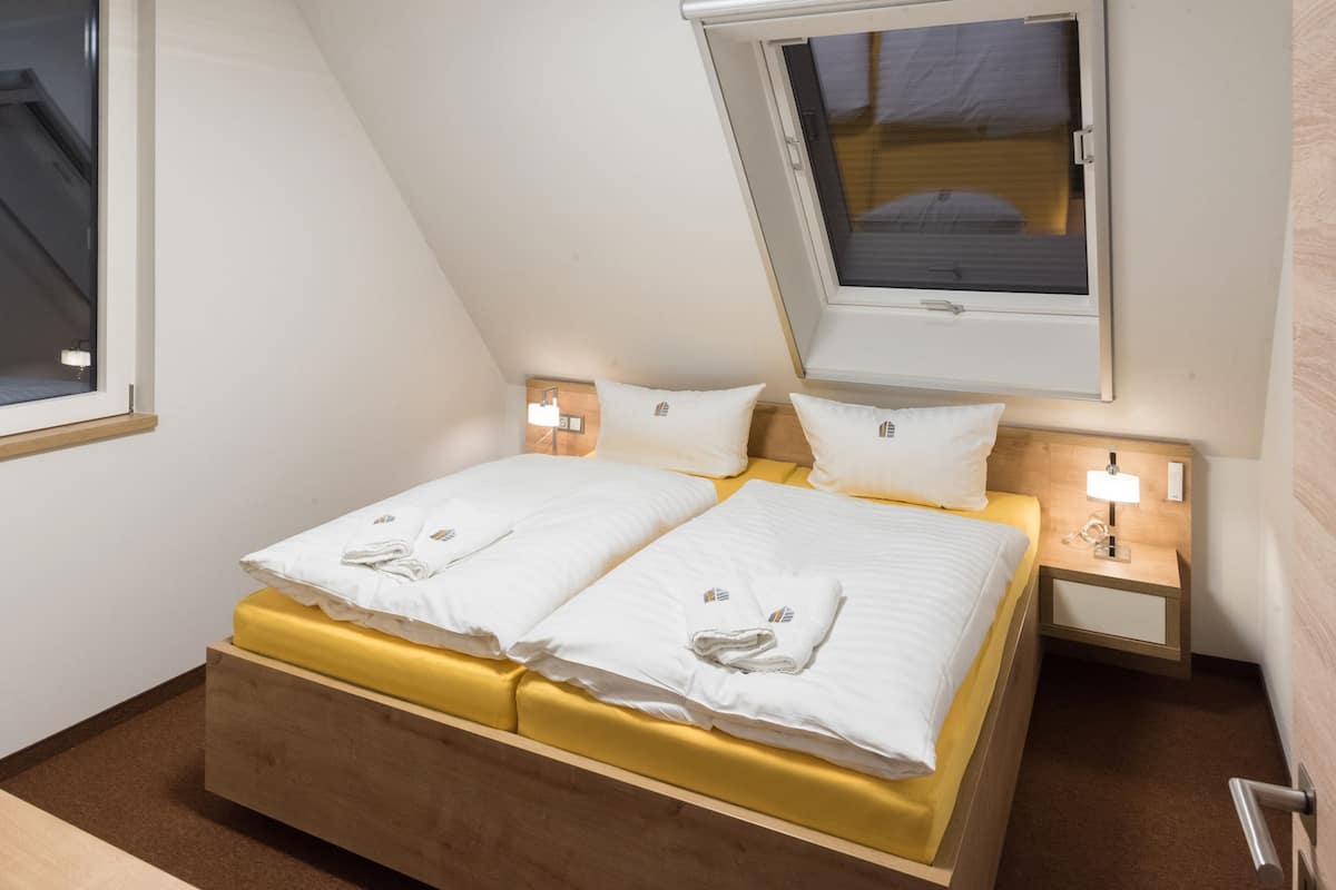 A neatly made double bed with white linens and a yellow headboard, a bedside table with a lamp, and a window with a view of the outside.