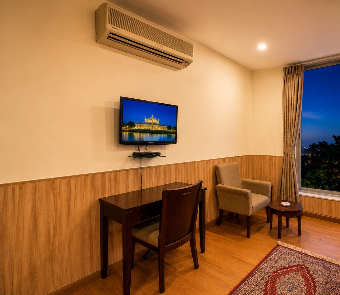 Una camera d'albergo con televisore a parete, una scrivania in legno, due sedie e una finestra con tende.