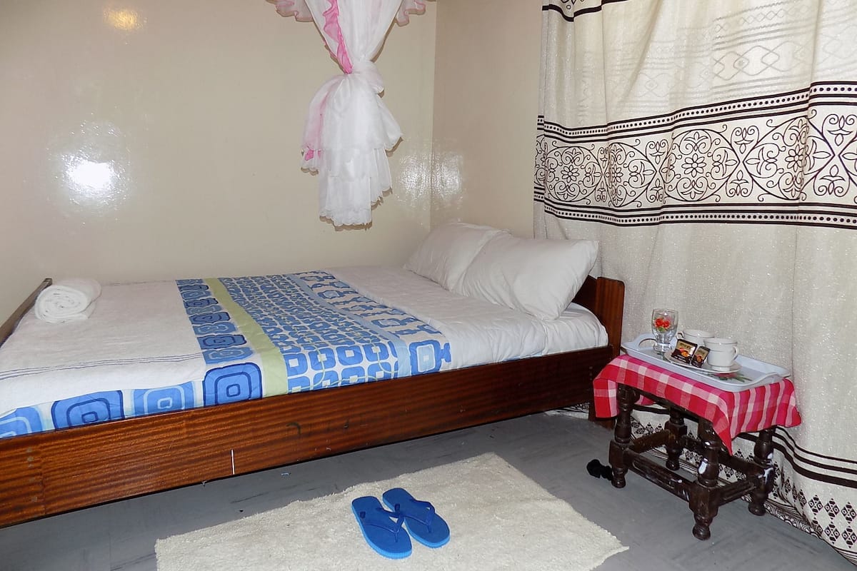 A bedroom with a bed, a bedside table with a red and white checkered tablecloth, a small wooden chair, and a pair of blue flip-flops on the floor.