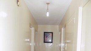 Un pasillo con paredes color beige, piso de baldosas y una lámpara en el techo. Se observa un cuadro enmarcado en la pared y una puerta a la derecha.