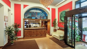 A lobby with a reception desk, a large arched window, a chandelier, a white sofa, and potted plants. - Hotel Oxford (Rome)