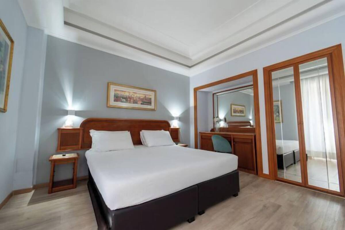 A hotel room with a large bed, bedside tables, a desk, a chair, a mirror, and a wardrobe.